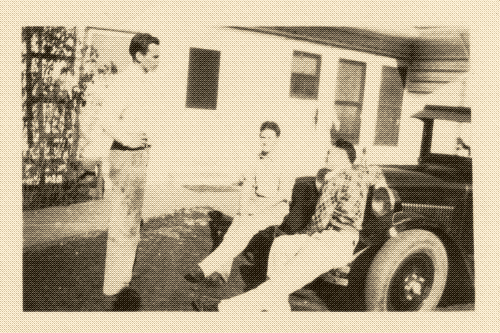 black and white photo of three men chatting while two of them sit on the hood of a car