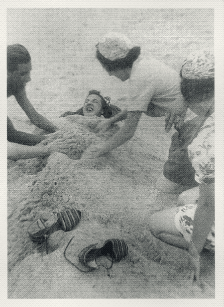black and white photo of a group of friends burying a laughing woman in sand on the beach